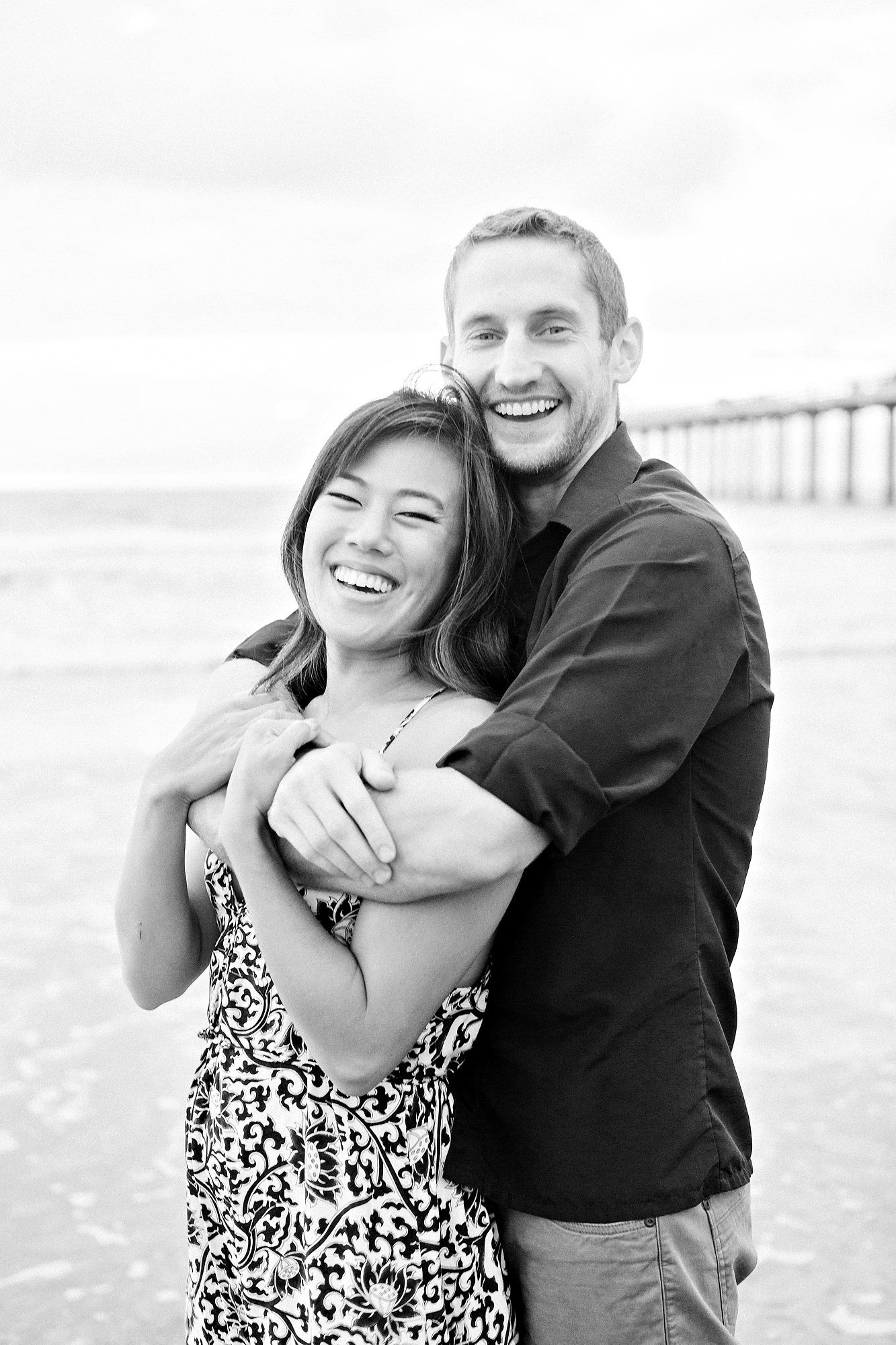 A smiling couple embraces on a beach with a pier in the background.