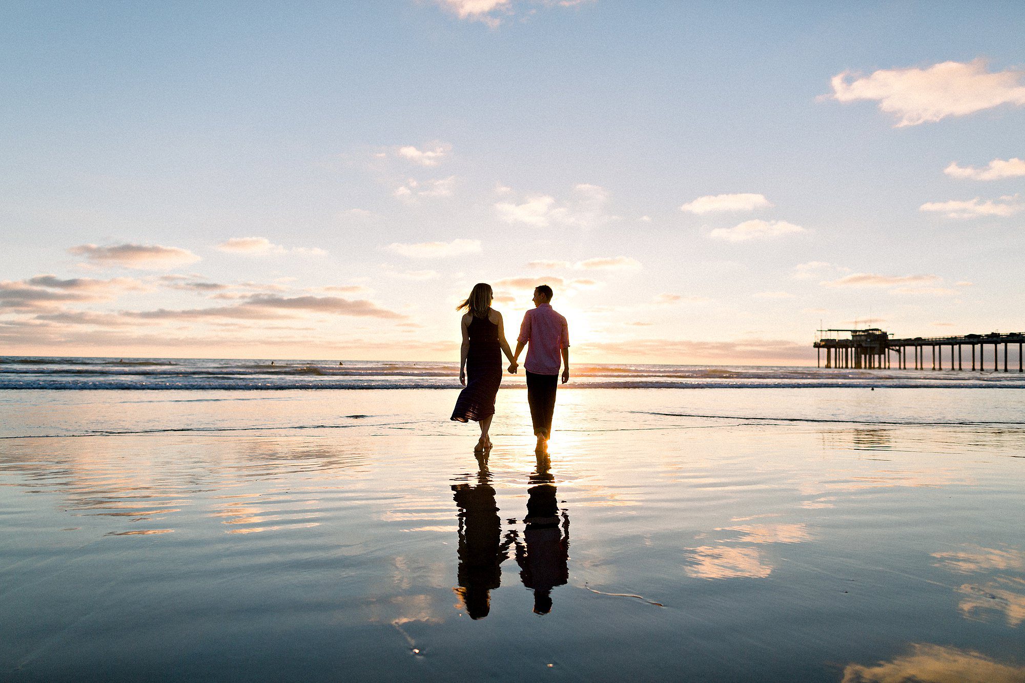 A couple holding hands walks along the beach at sunset, with reflections in the wet sand and a pier in the distance.