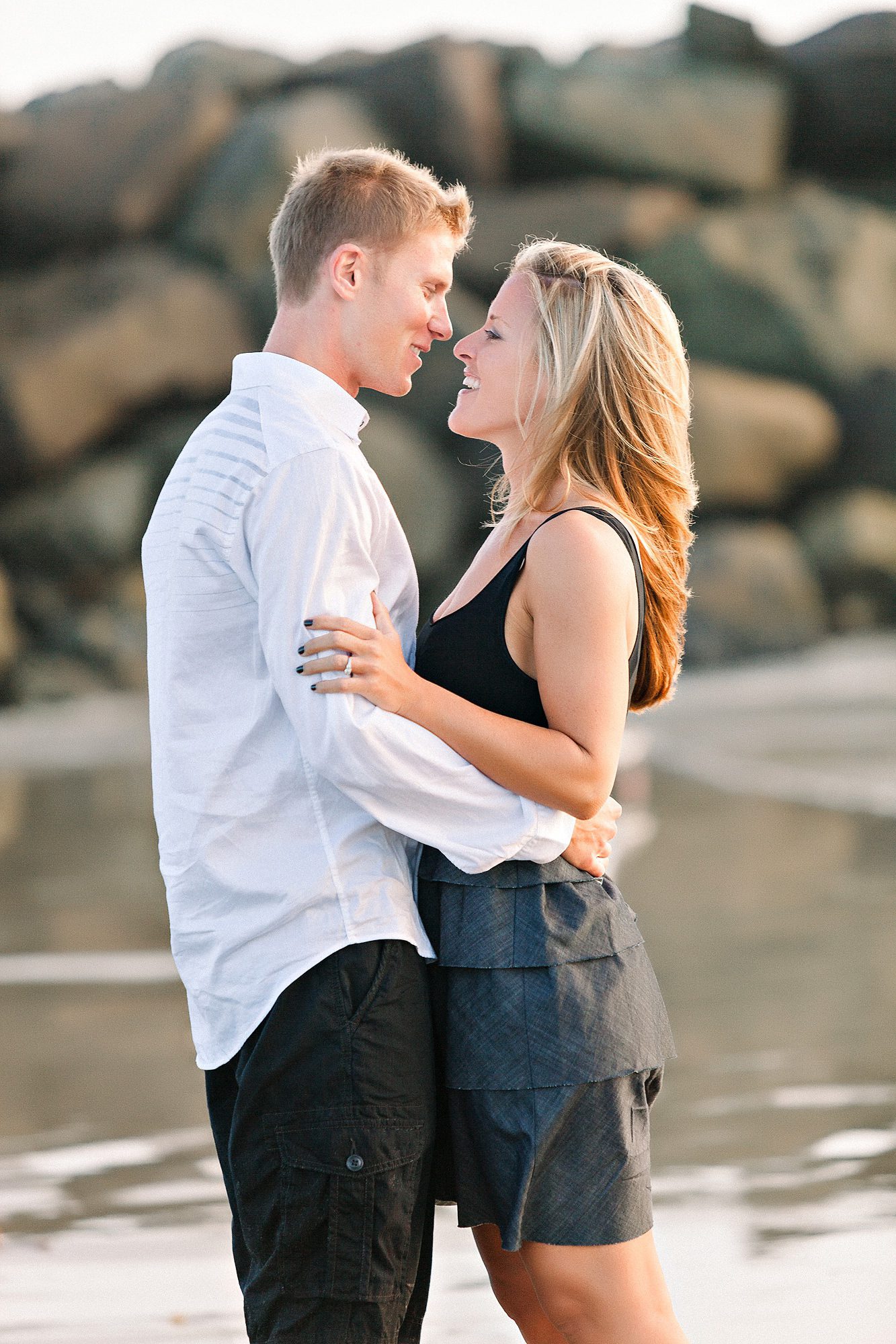 A couple stands close together on a beach, smiling at each other with rocks in the background.