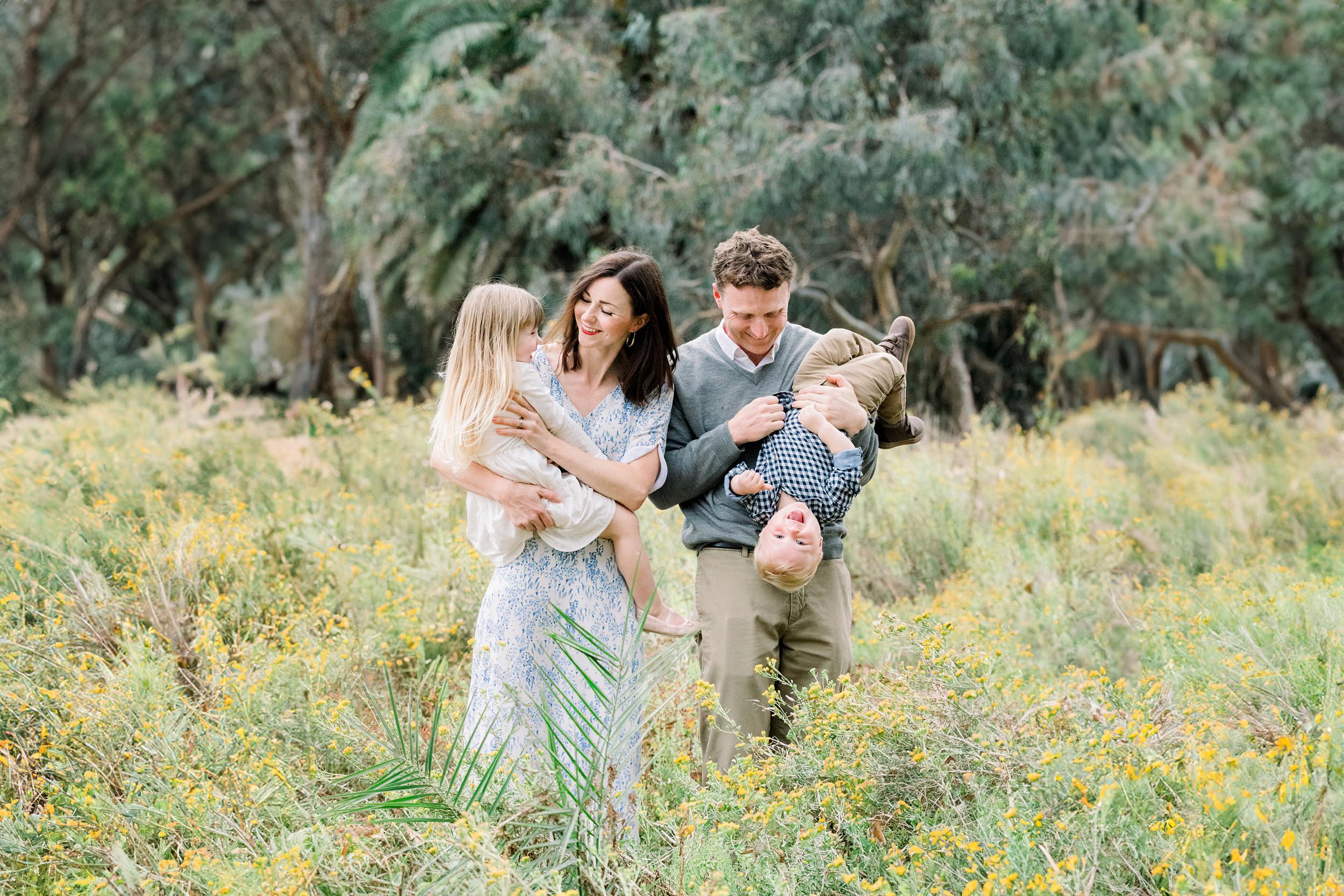 A family of four smiles and plays together in a field of yellow wildflowers surrounded by trees.