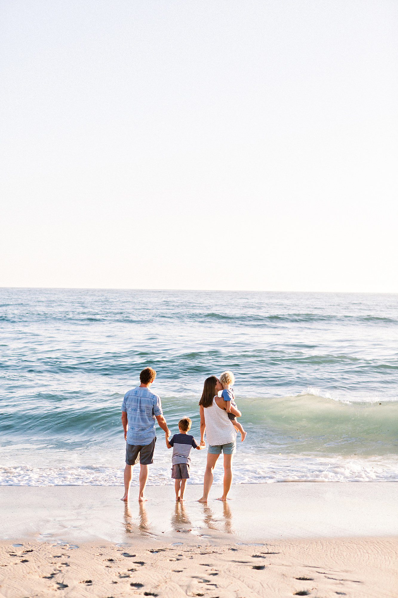 A family of four stands at the edge of the ocean, holding hands and looking out at the waves on a sunny day.