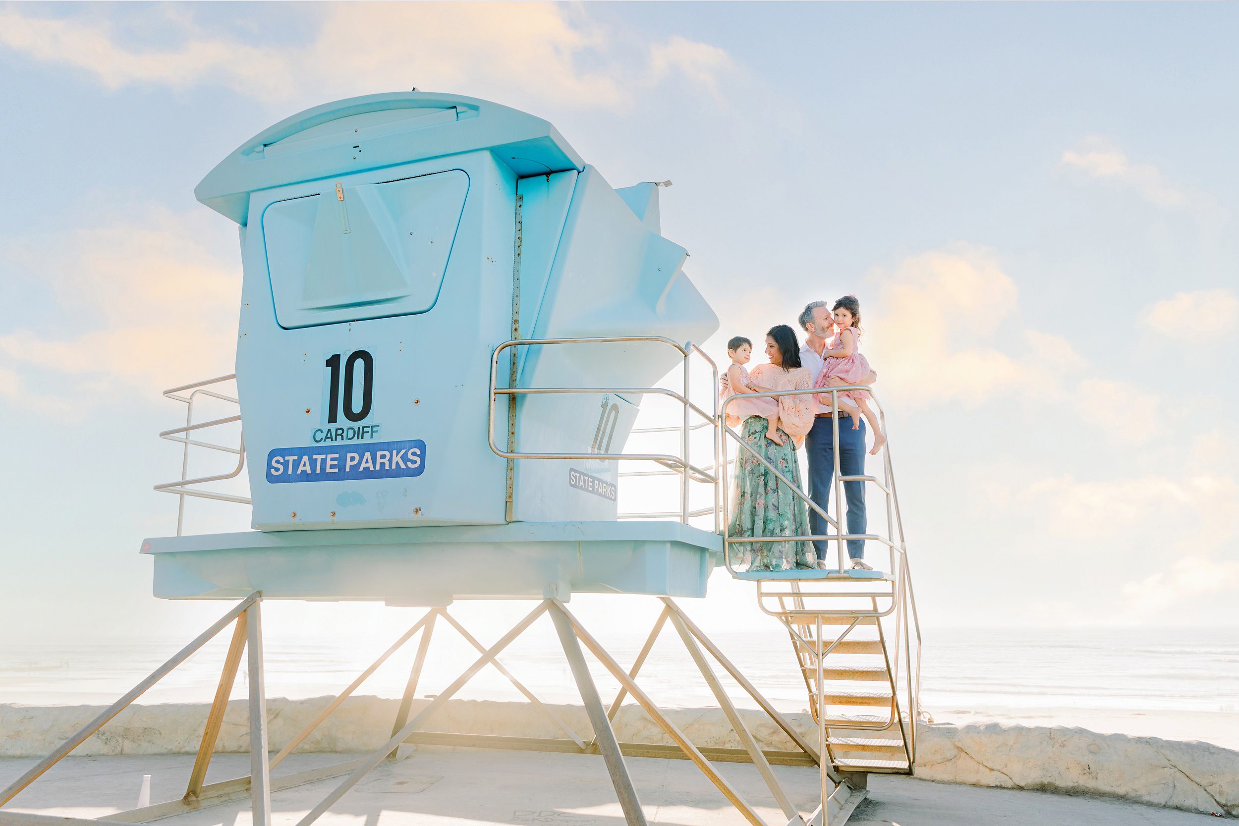 Four people stand on the stairs of a light blue lifeguard tower by the beach under a pastel sky.