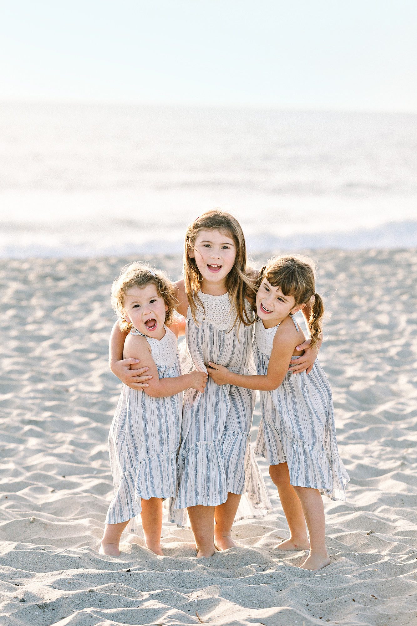 Three young girls in striped dresses smile and hug while standing barefoot on a sandy beach with the ocean behind them.