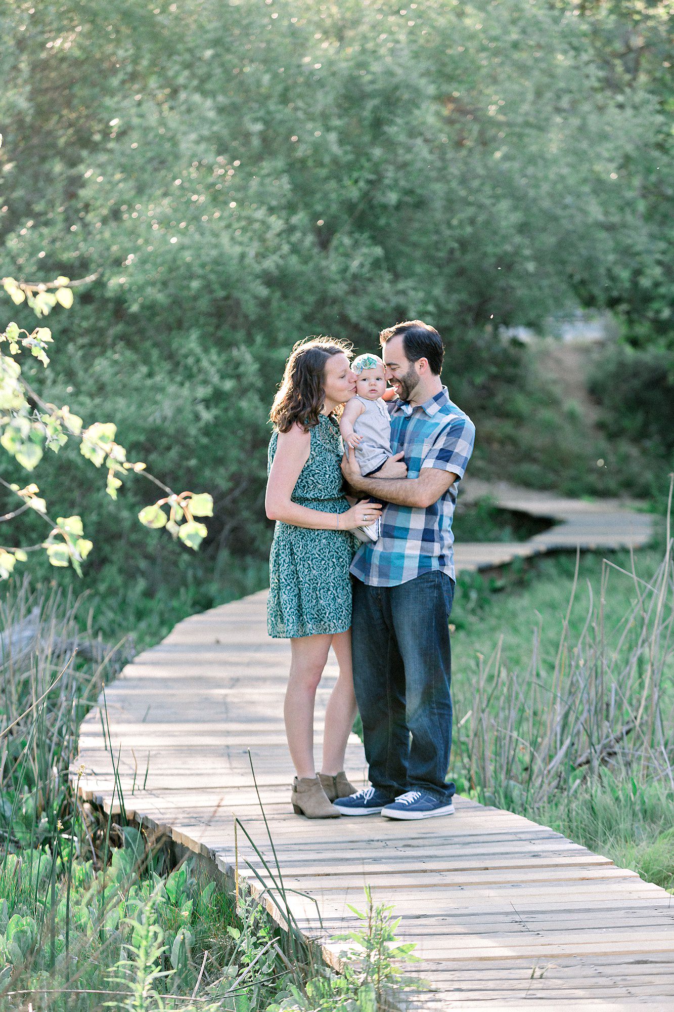 A couple stands on a wooden path in a green park, holding their baby and smiling at each other.