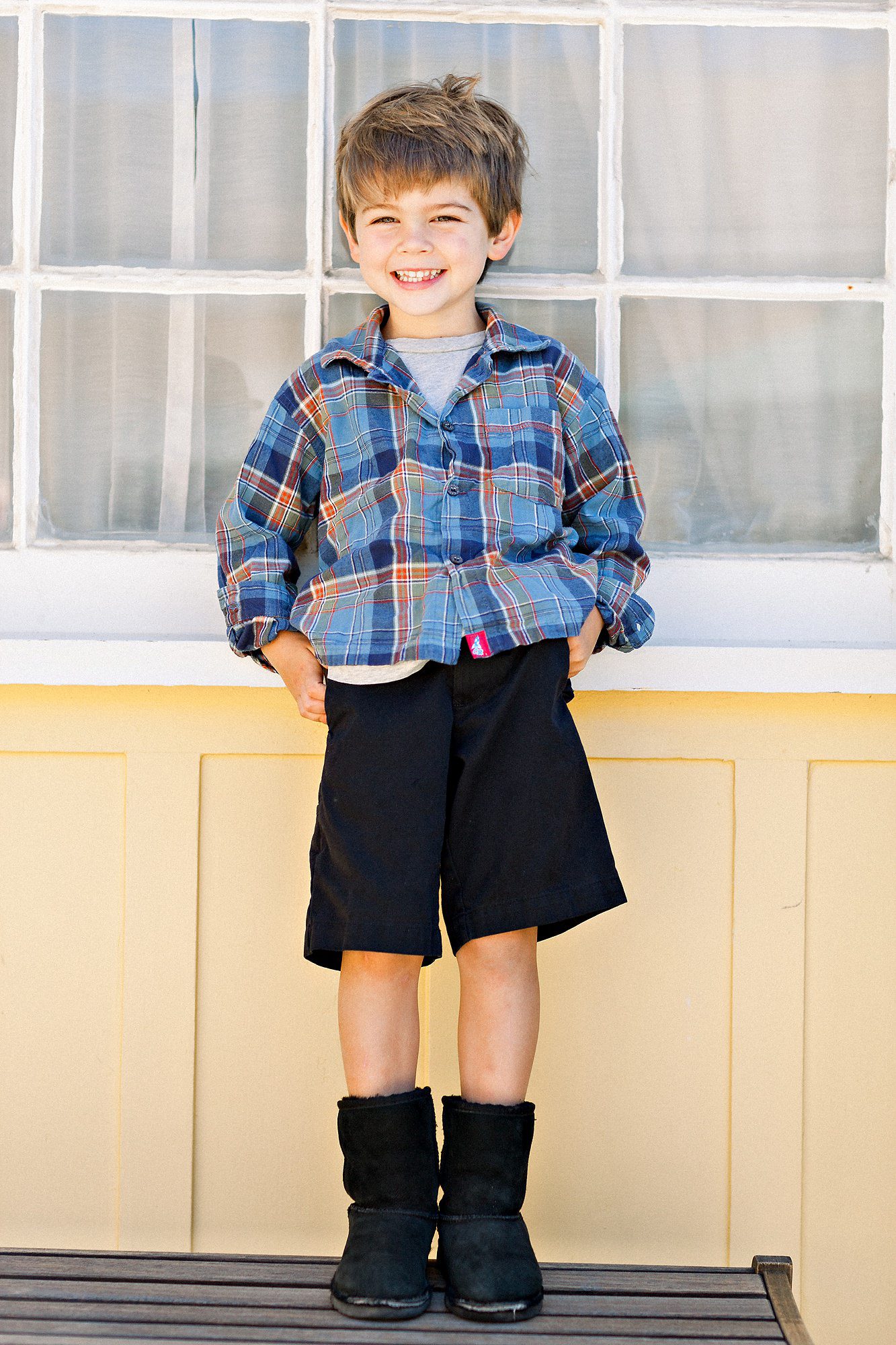 Smiling young boy in plaid shirt, black shorts, and boots stands in front of a window with hands in his pockets.