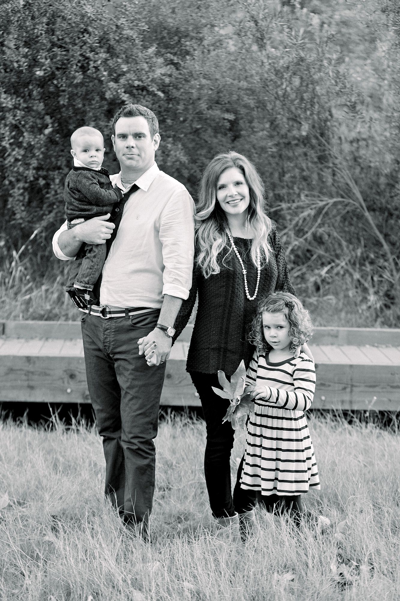 A family of four poses outdoors; parents stand with a baby and a young girl in striped dress holding leaves.