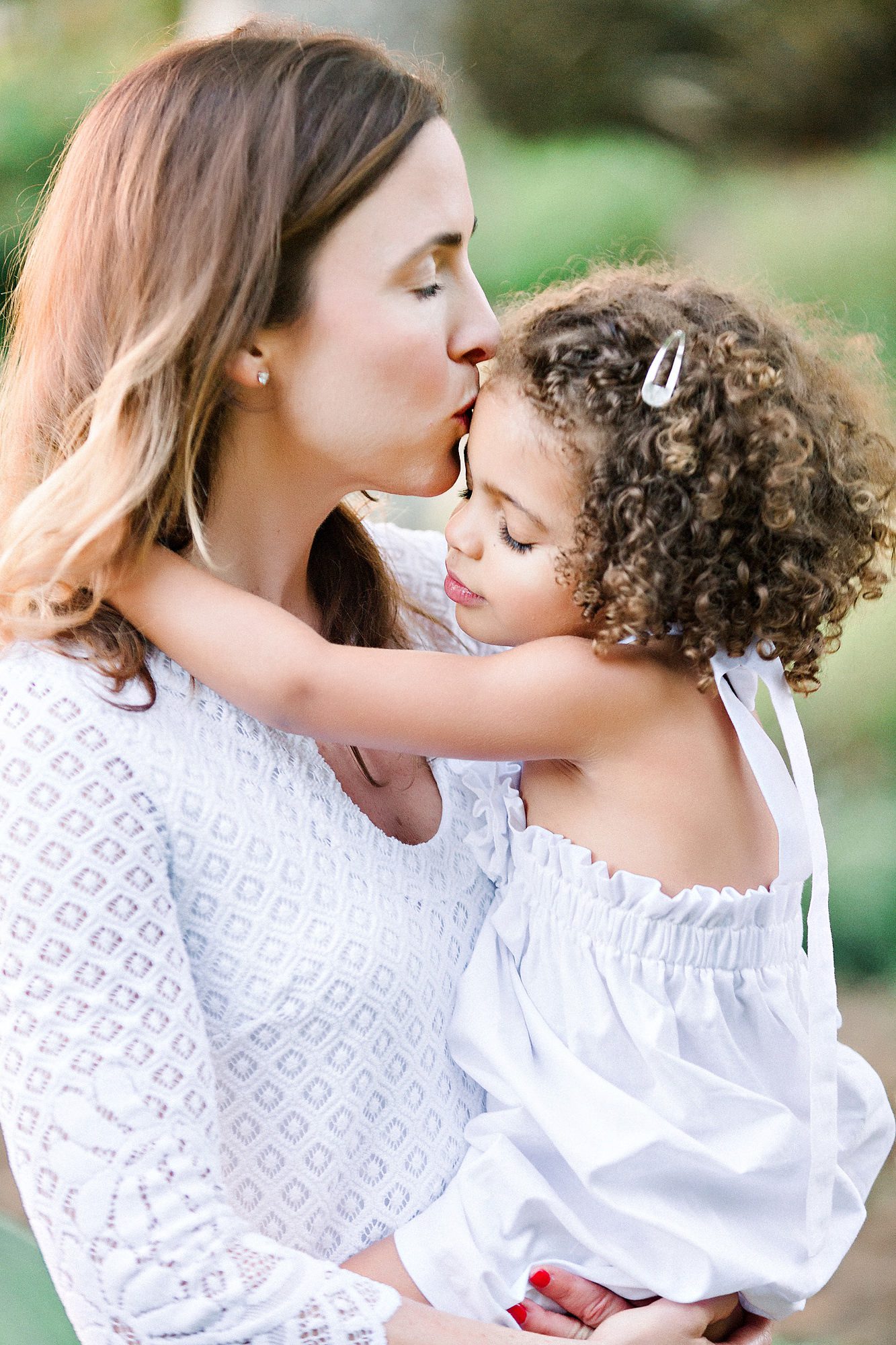 Woman kisses young girl with curly hair on the forehead while hugging her outdoors, both dressed in white.