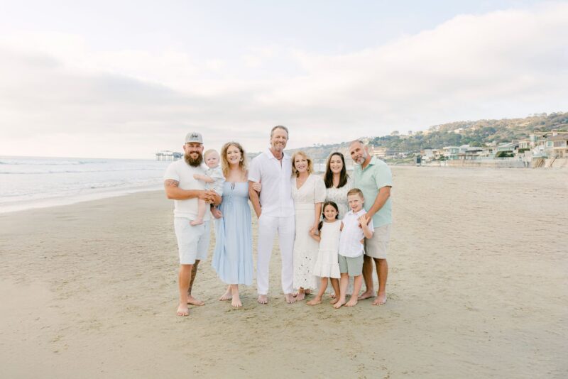 Three adults, kids, and a toddler pose on a sandy beach with houses—captured by Carlsbad photographers.