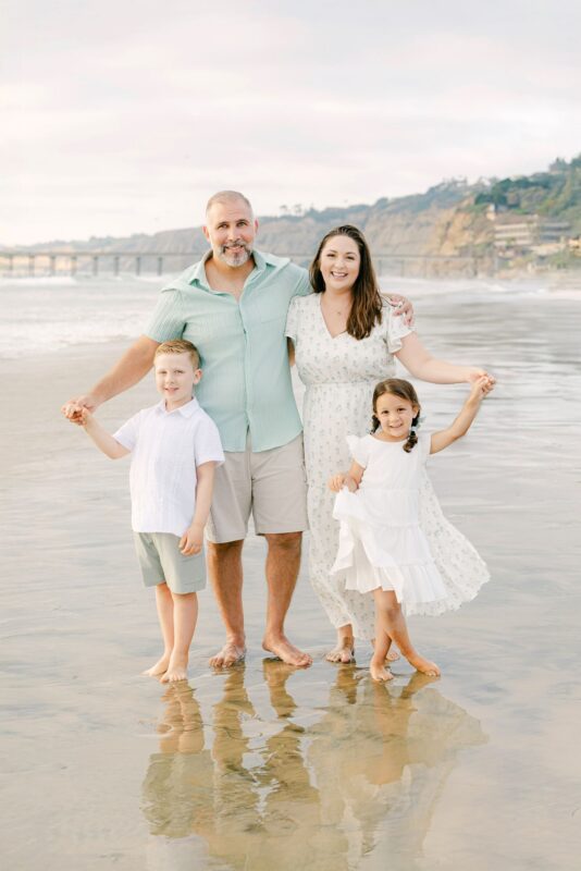 Smiling family of four holding hands, barefoot on a sandy beach—captured by talented Carlsbad family photographers.