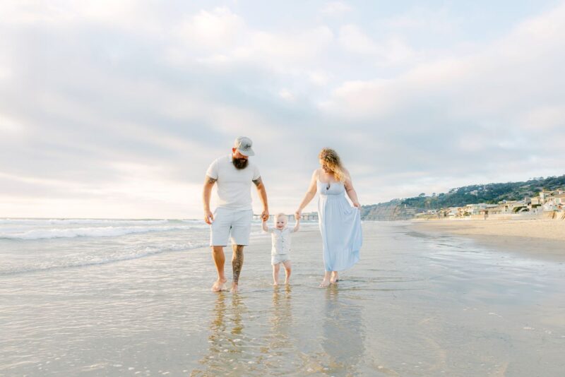A family of three walks barefoot on a sandy beach, captured by family photographers with the ocean and houses behind.