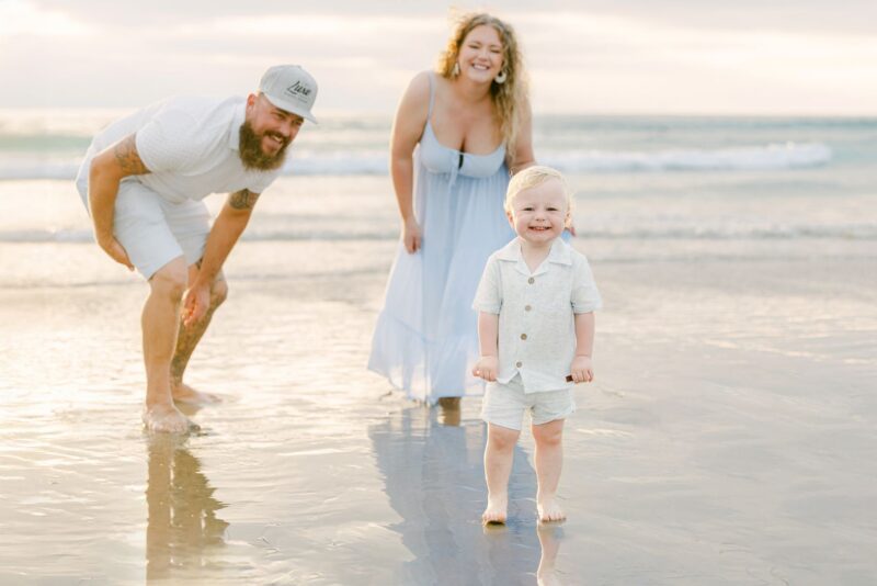 A smiling toddler stands on a beach with two happy adults, captured by family photographers, all barefoot by the water’s edge.
