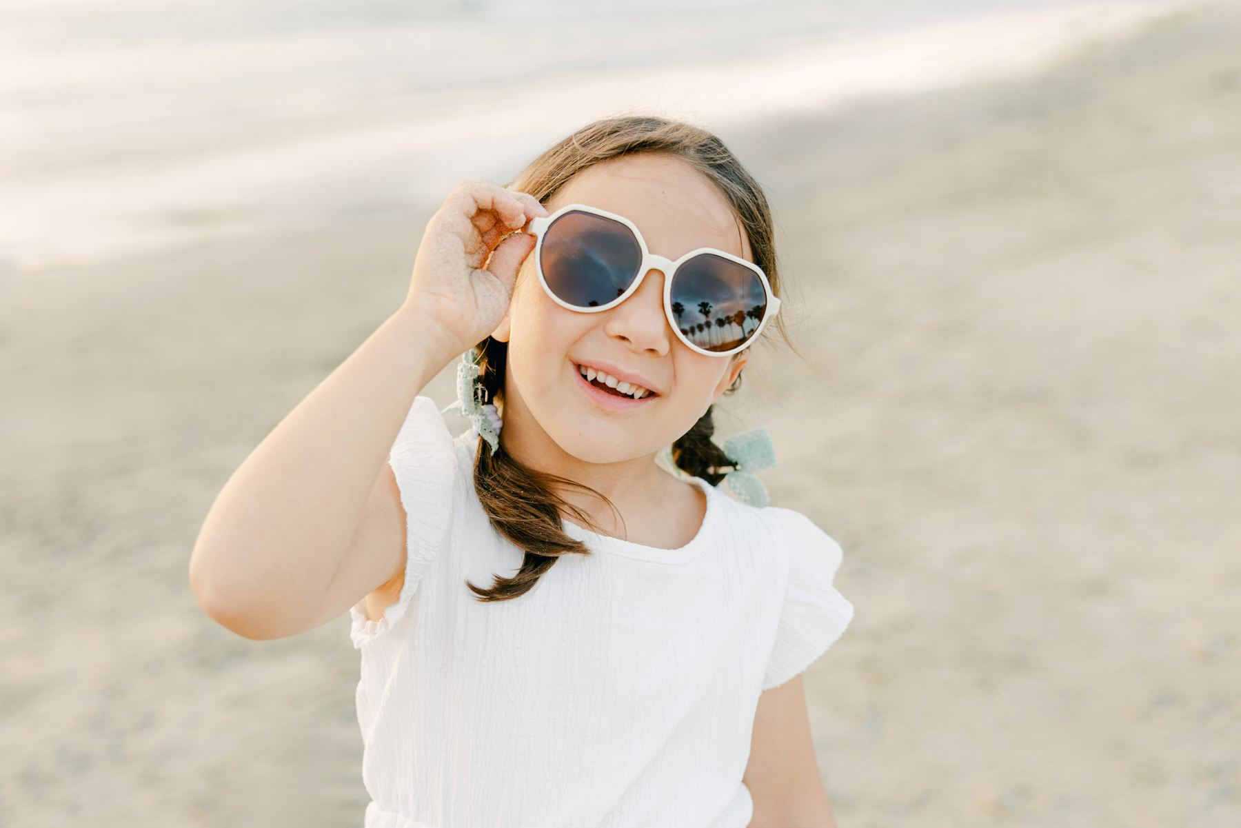 Smiling child with braided hair and sunglasses stands on a sunny beach, captured by Carlsbad photographers.