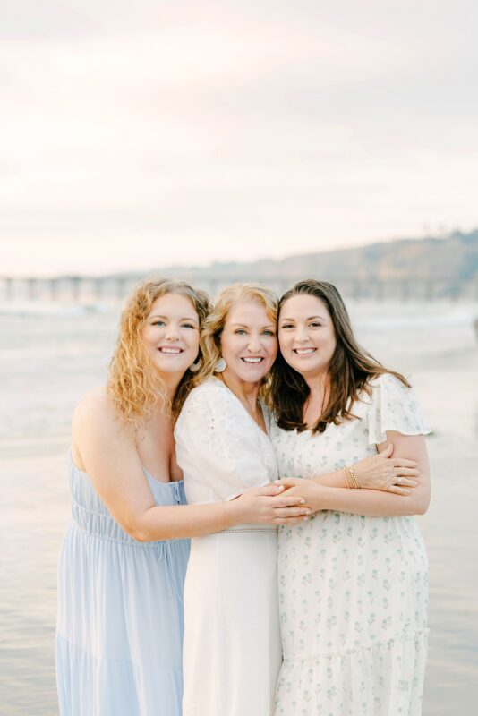 Three women in light dresses smile and hug on a beach—captured by a talented Carlsbad family photographer.