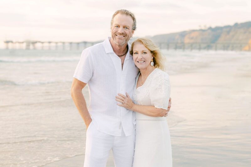 Smiling couple in white stands on a beach near a pier and cliffs—perfect inspiration for family photographers in Carlsbad.