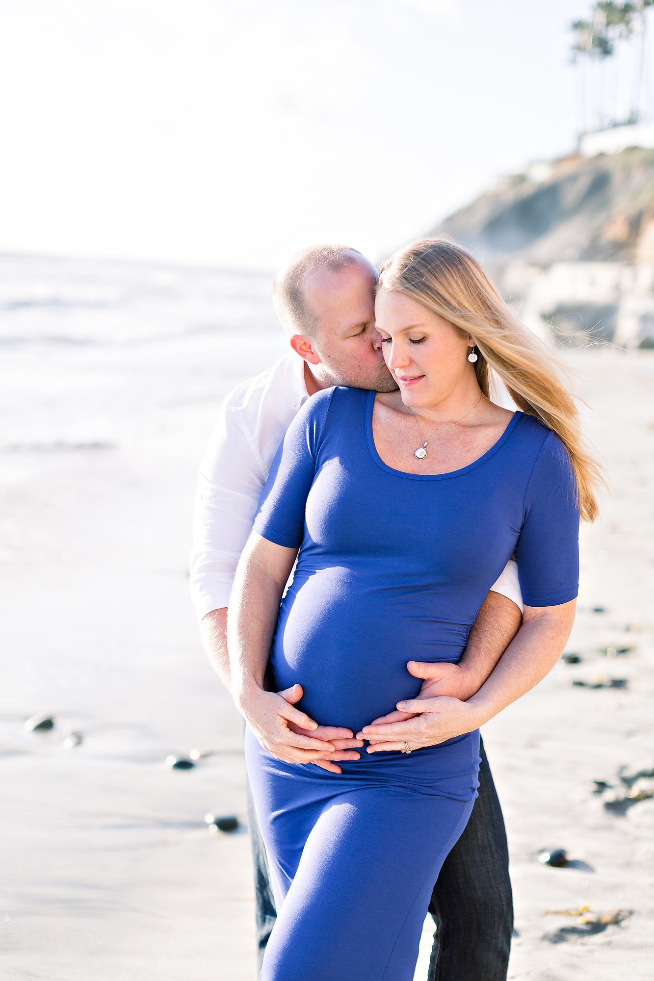 A man embraces and kisses a pregnant woman in a blue dress on a beach, both looking serene.