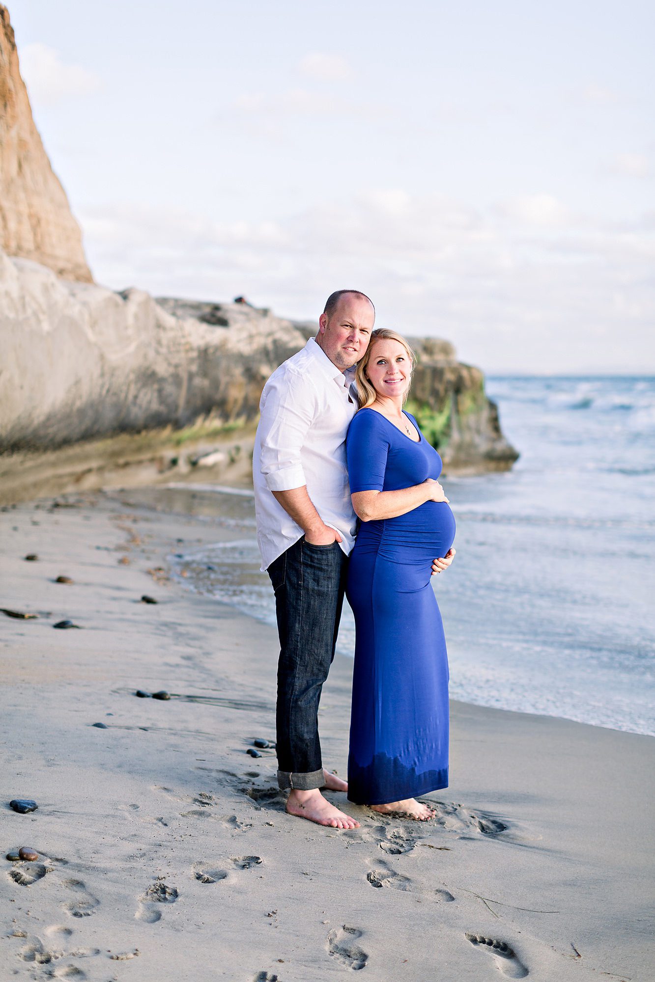 A smiling couple stands on a beach, the woman in a blue dress cradling her pregnant belly, with cliffs in the background.