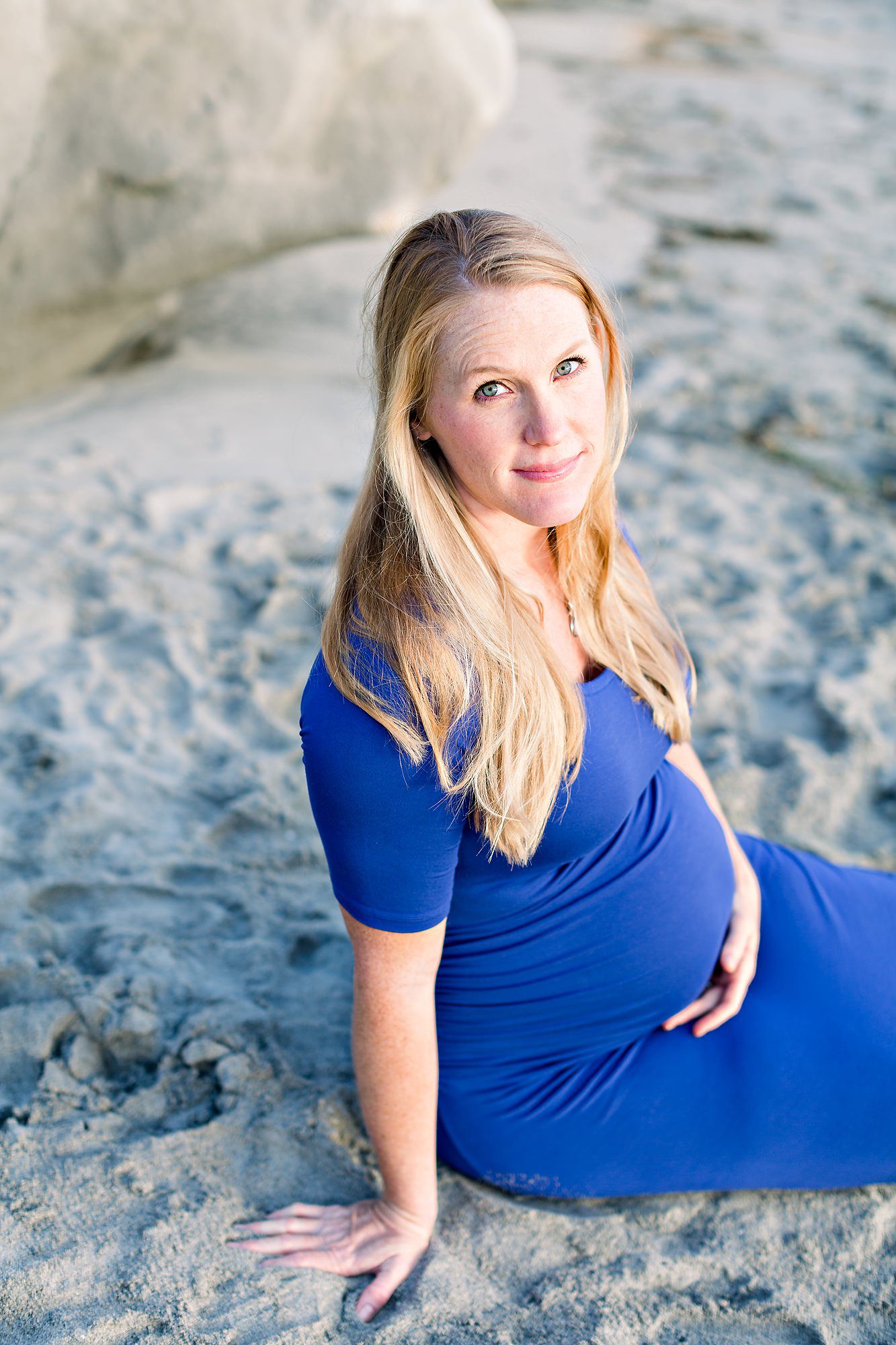 Pregnant woman in a blue dress sits on sandy beach, looking up and smiling gently at the camera.