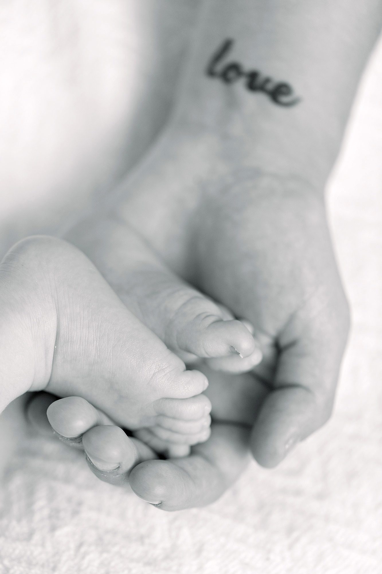 A babys foot rests gently in an adults hand with a love tattoo on the wrist.