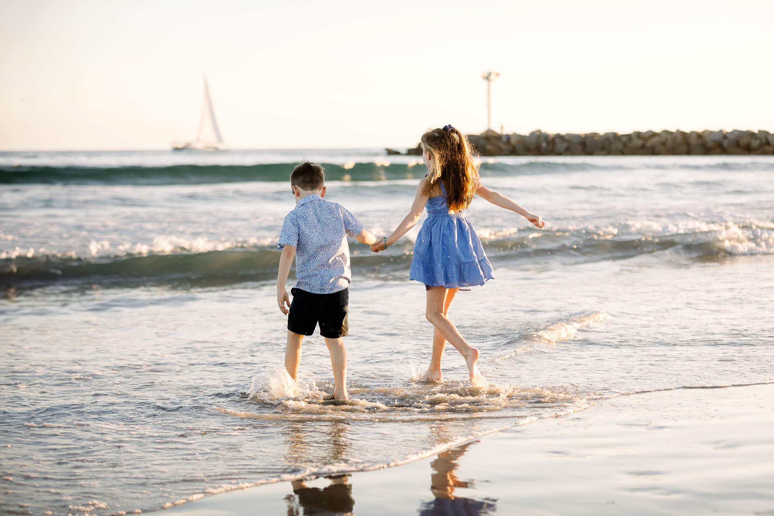 Two children holding hands walk in shallow water at Carlsbad Beach, perfect for family portraits with a sailboat behind.