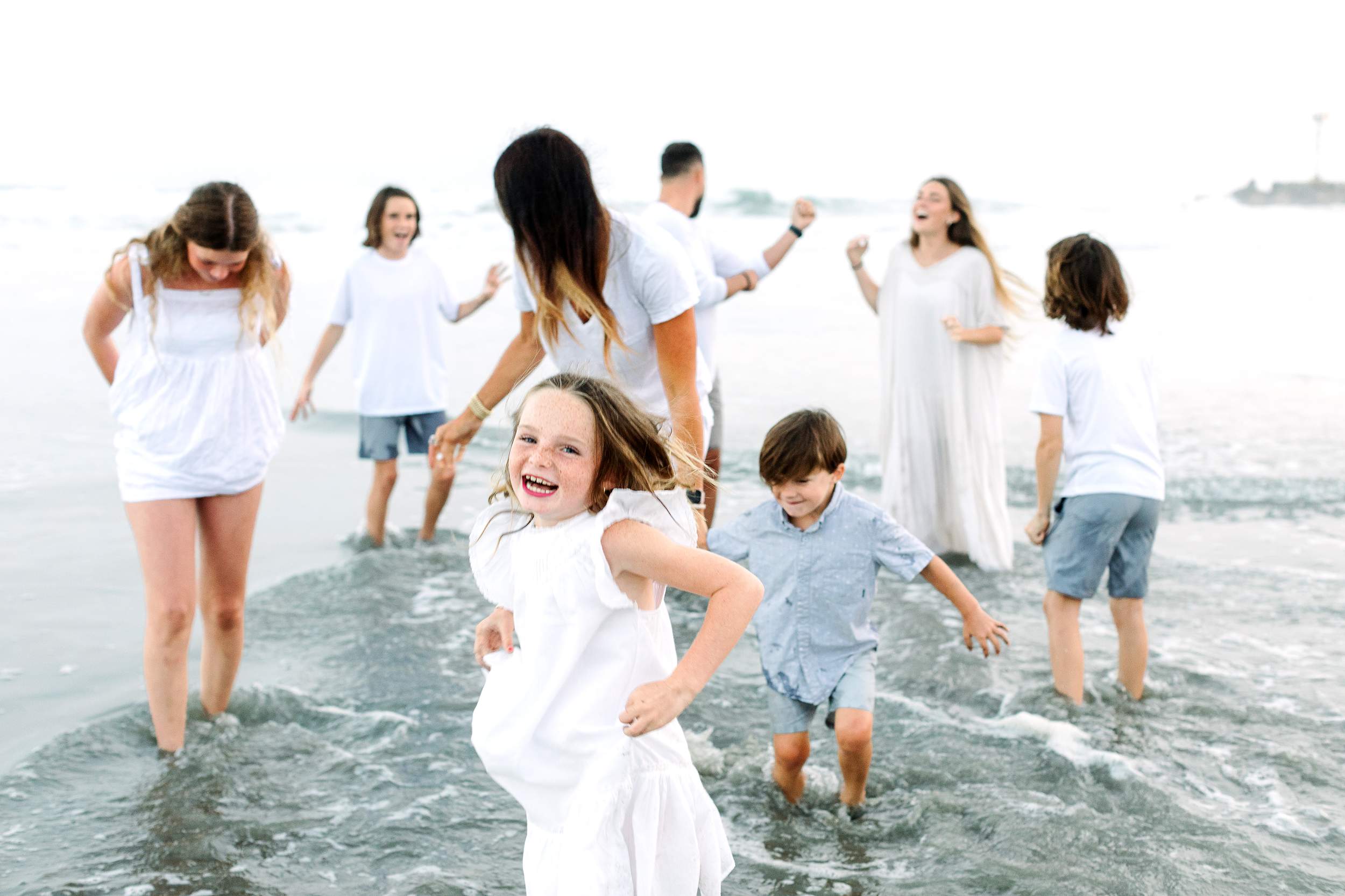 A group laughs and plays in shallow ocean water during Carlsbad Beach family portraits.