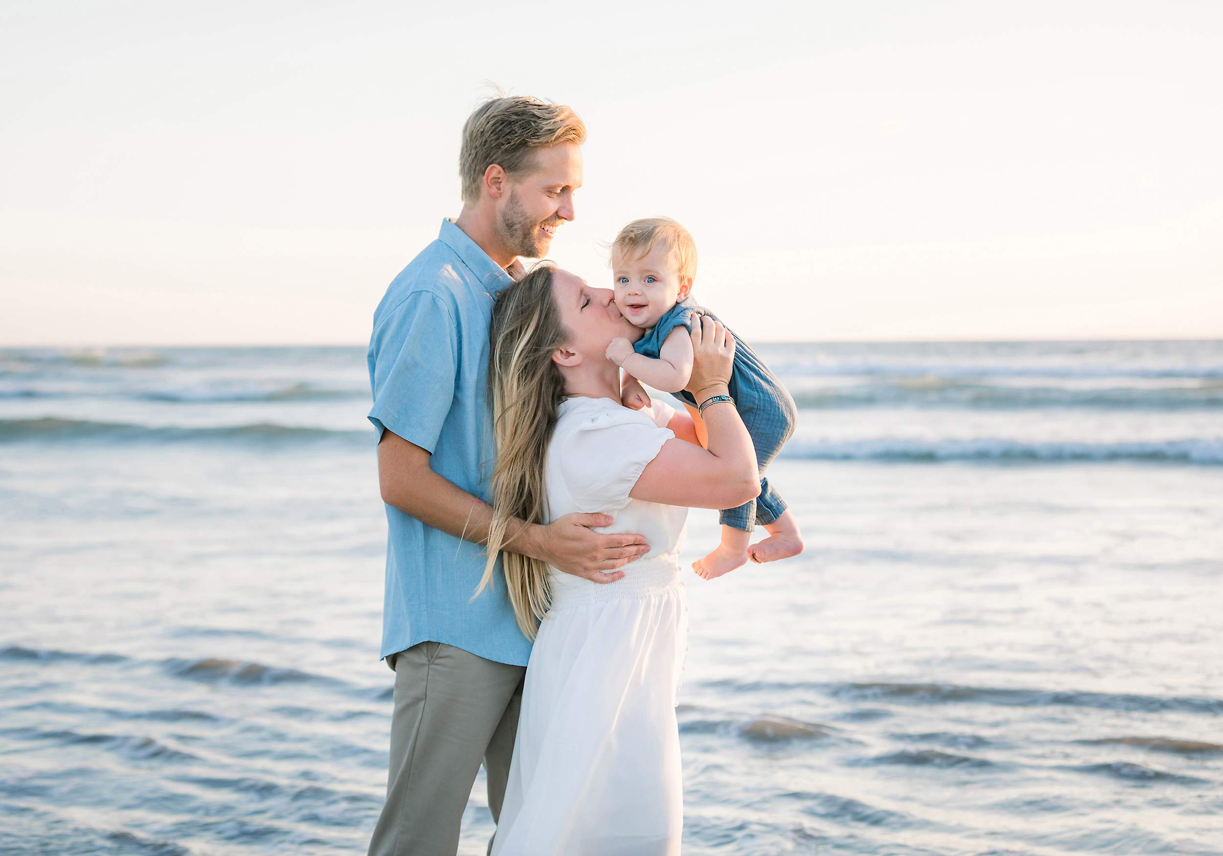 A smiling couple holds their laughing baby at Carlsbad Beach with ocean waves in the background—perfect family portraits.