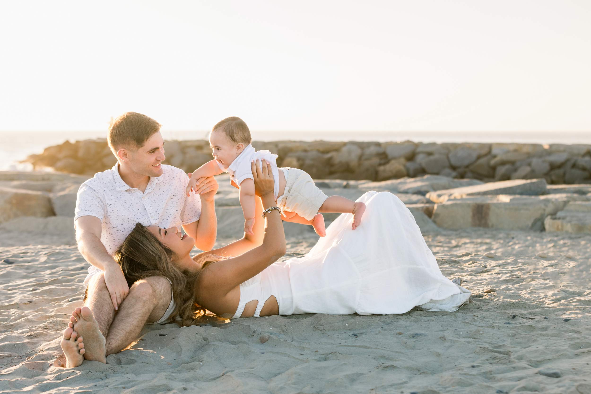 A couple enjoys Carlsbad Beach family portraits at sunset, smiling as the woman lifts their baby against the glowing sky.