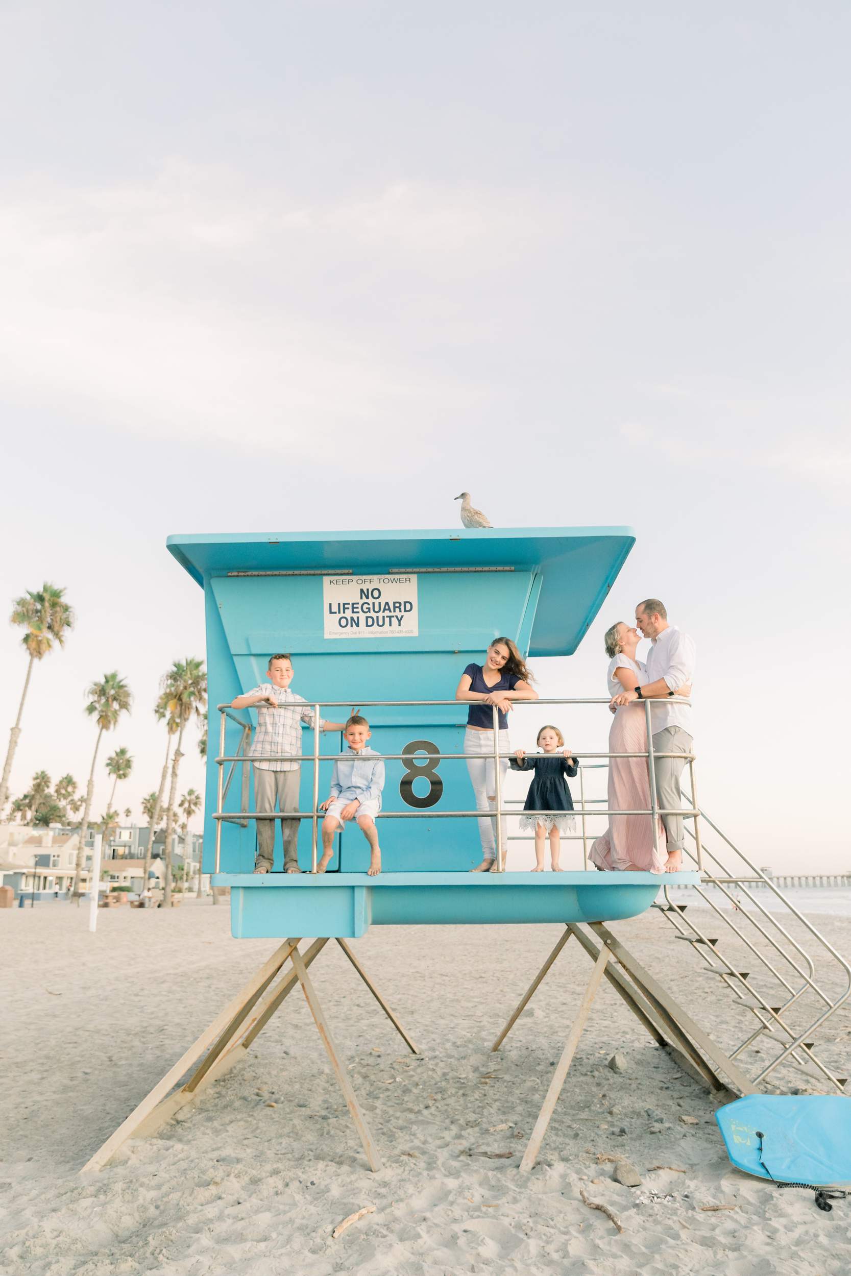 A family of six poses on a blue lifeguard tower at Carlsbad Beach, with palm trees and a seagull in the background.