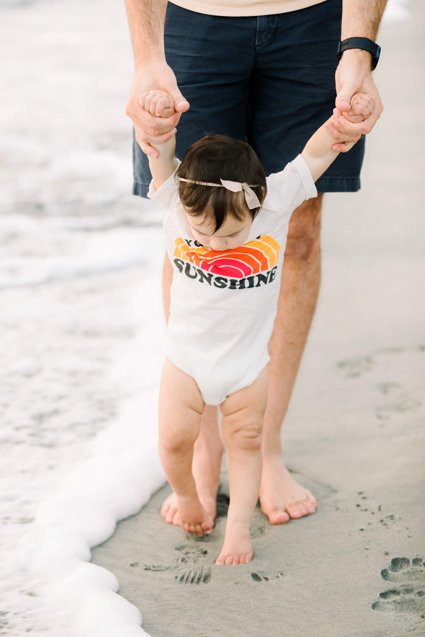 Adult helping a baby walk near the ocean, holding hands; Carlsbad Beach Family Portraits capture sweet sunshine moments.