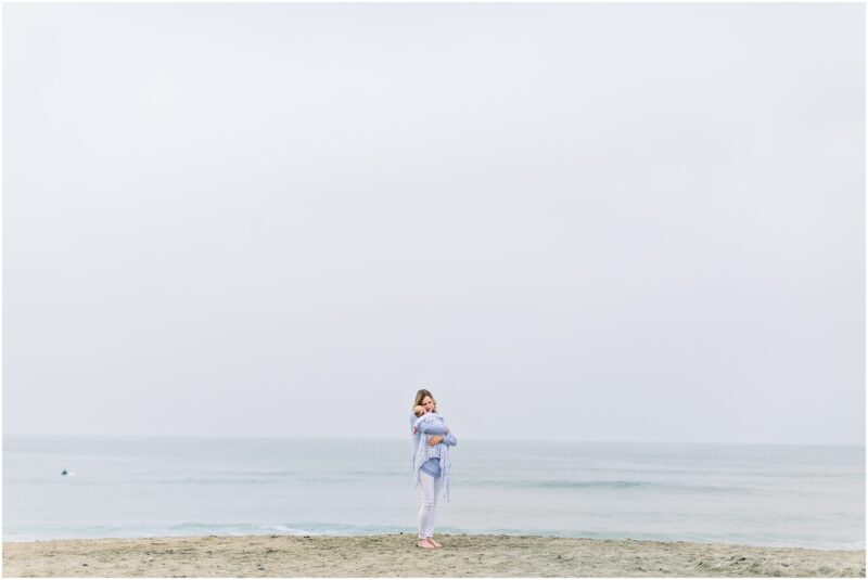 A woman holding a baby on a beach, captured by a Carlsbad Newborn Photographer.