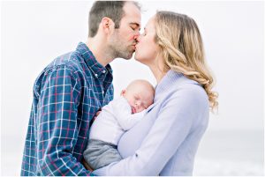 A couple kisses while holding a sleeping baby, wrapped in cozy clothing. Carlsbad Newborn Photographer.