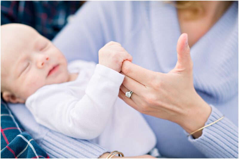 A baby in white sleeps, gently holding an adult’s thumb with a diamond ring. Carlsbad Newborn Photographer.