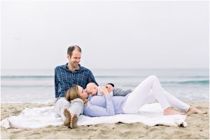A couple relaxes on a beach blanket, the woman holding her baby—captured by a Carlsbad Newborn Photographer.