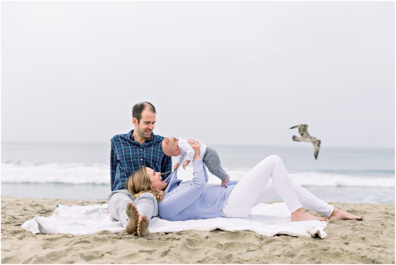 A family with a baby relaxes on a blanket at the beach—captured by your Carlsbad Newborn Photographer.