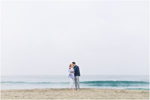 A couple stands close together on a sandy beach under a cloudy sky, captured by a Carlsbad Newborn Photographer.