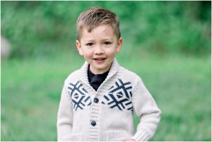 Young boy in a cream sweater with black patterns smiles during Solana Beach pregnancy photos on the grass.