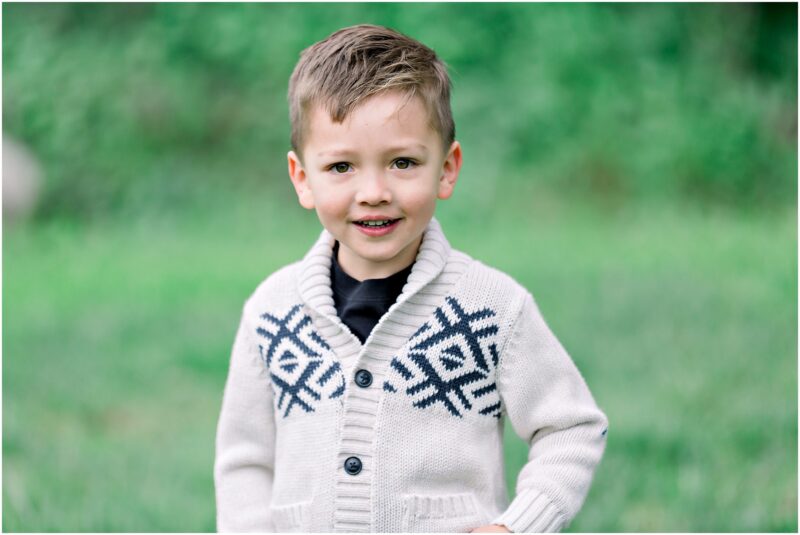 Young boy in a cream sweater with black patterns smiles during Solana Beach pregnancy photos on the grass.