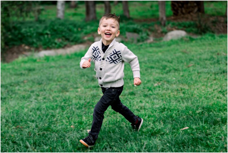 Young boy in a white sweater and black pants running joyfully during a Solana Beach maternity session in a park.