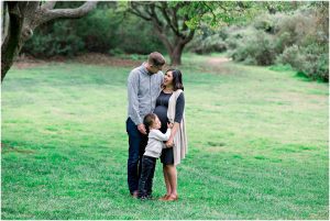 A family of three smiles and holds hands under a tree during a Solana Beach pregnancy photos session.