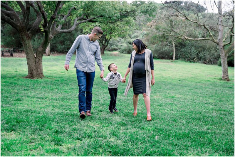 A family of three enjoys green grass in a park, smiling during their Solana Beach pregnancy photos session.