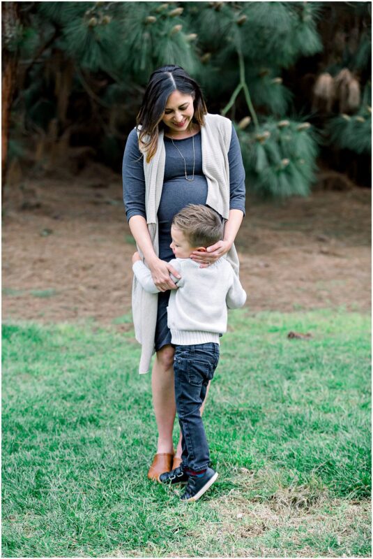 A pregnant woman smiles as a young boy hugs her waist during a sweet maternity session on grass with trees behind them.