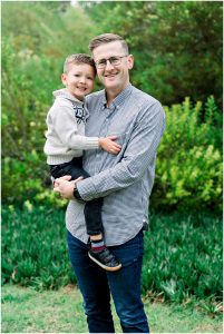 A smiling man holds a young boy in his arms, both standing outside in a garden during a Solana Beach pregnancy photos session.