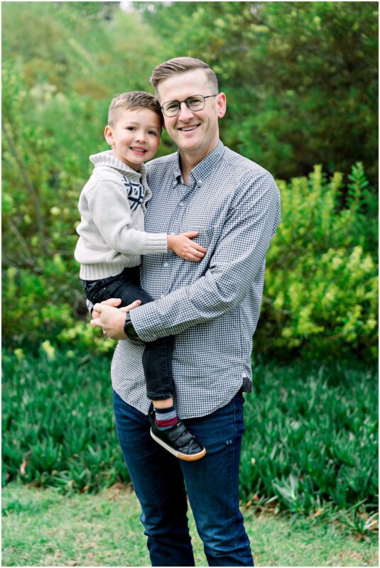 A smiling man holds a young boy in his arms, both standing outside in a garden during a Solana Beach pregnancy photos session.