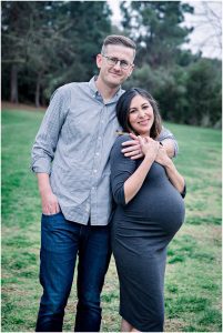 A smiling couple enjoys their Solana Beach pregnancy photos, with her in a gray dress and him in a checkered shirt.