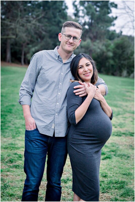 A smiling couple enjoys their Solana Beach pregnancy photos, with her in a gray dress and him in a checkered shirt.