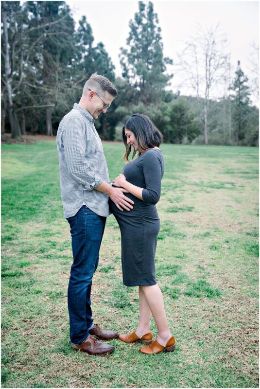 A man and a pregnant woman smile in a grassy field during their Solana Beach pregnancy photos maternity session.