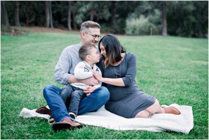 A family of three sits closely on a blanket in the park, smiling and cuddling during their Solana Beach pregnancy photos.
