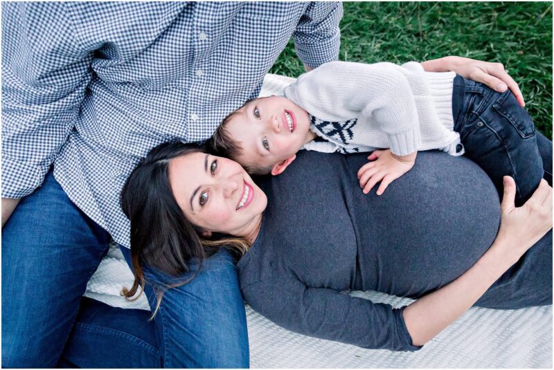 A pregnant woman smiles on a blanket as a child hugs her belly during a Solana Beach pregnancy photos maternity session.