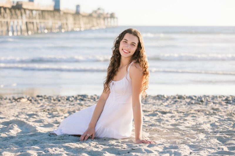 Smiling woman in a white dress sits on sandy beach with ocean and pier—perfect for beach senior photos.