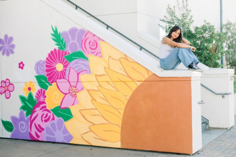 A high school senior poses on stairs by a vibrant flower mural, perfect for high school senior portraits.