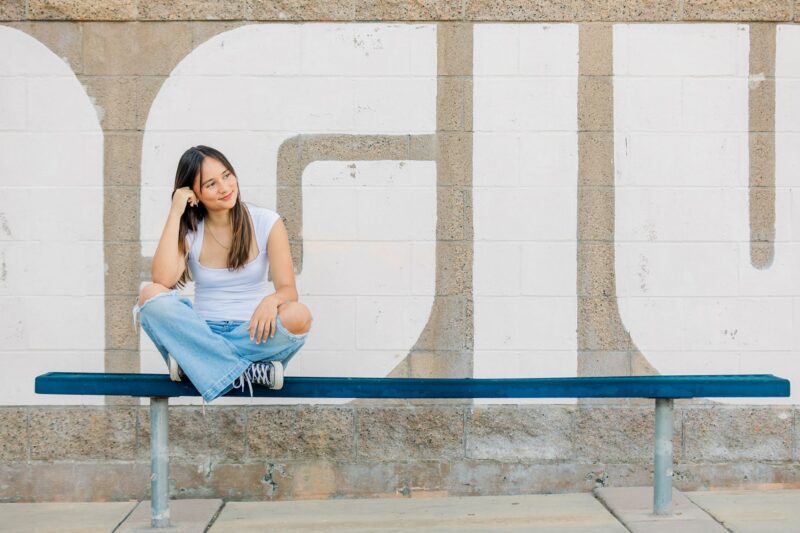 High school senior portrait: Young woman in jeans and a white top sits casually on a blue bench, smiling by a painted brick wall.